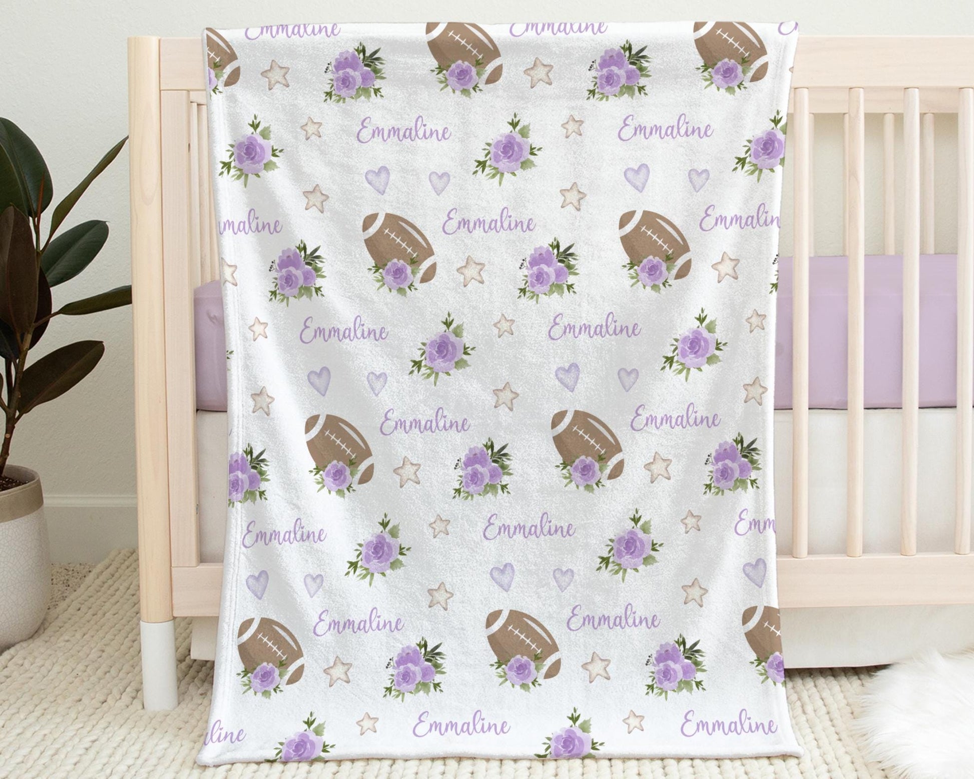 Baby blanket with footballs and purple flowers and personalized with baby's name in purple pictured hanging on the side of a crib.