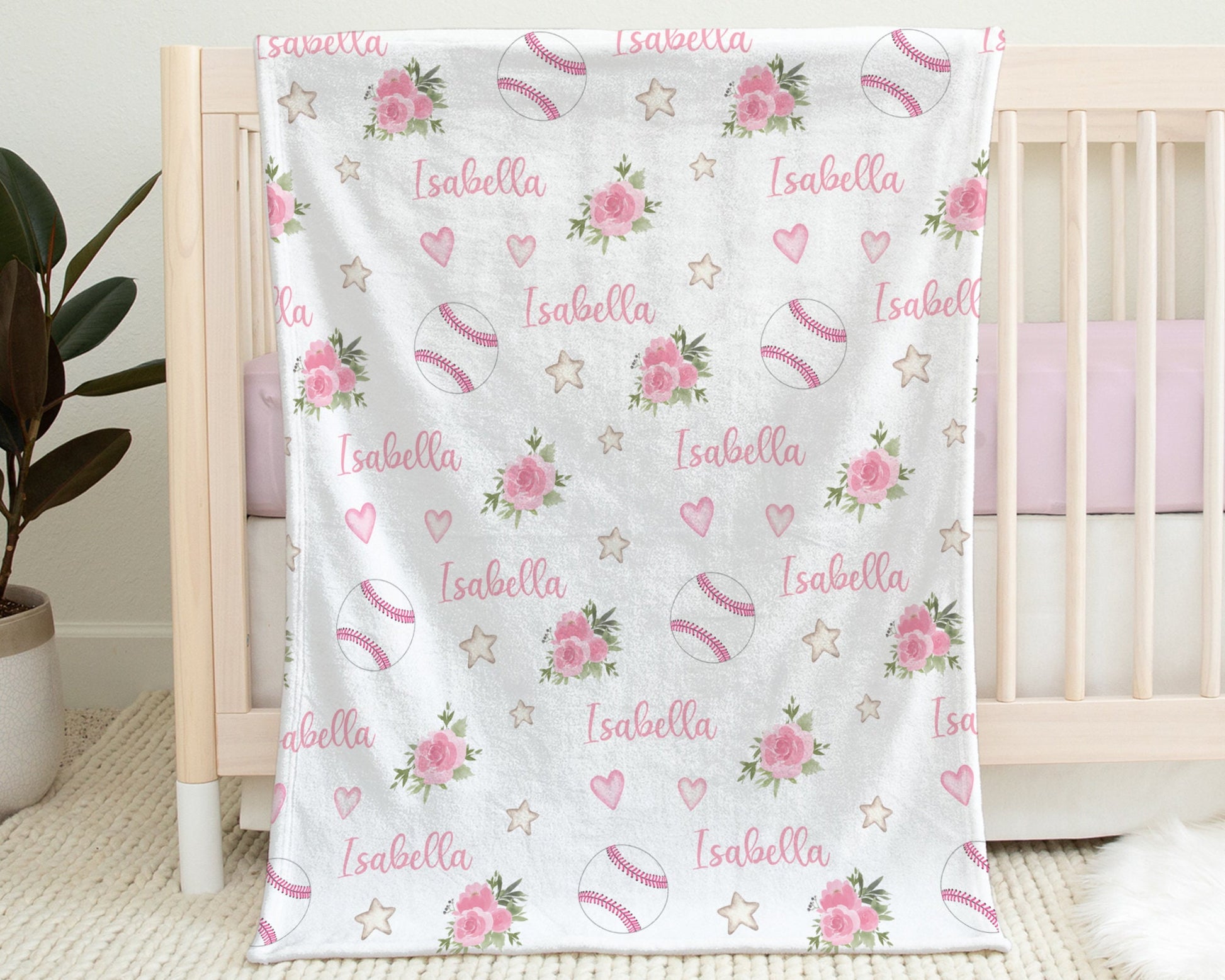 Baby blanket with baseballs with pink stitching and pink flowers and personalized with baby's name in pink pictured hanging on the side of a crib in baby's nursery.