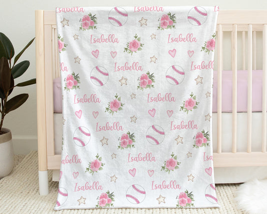 Baby blanket with baseballs with pink stitching and pink flowers and personalized with baby's name in pink pictured hanging on the side of a crib in baby's nursery.