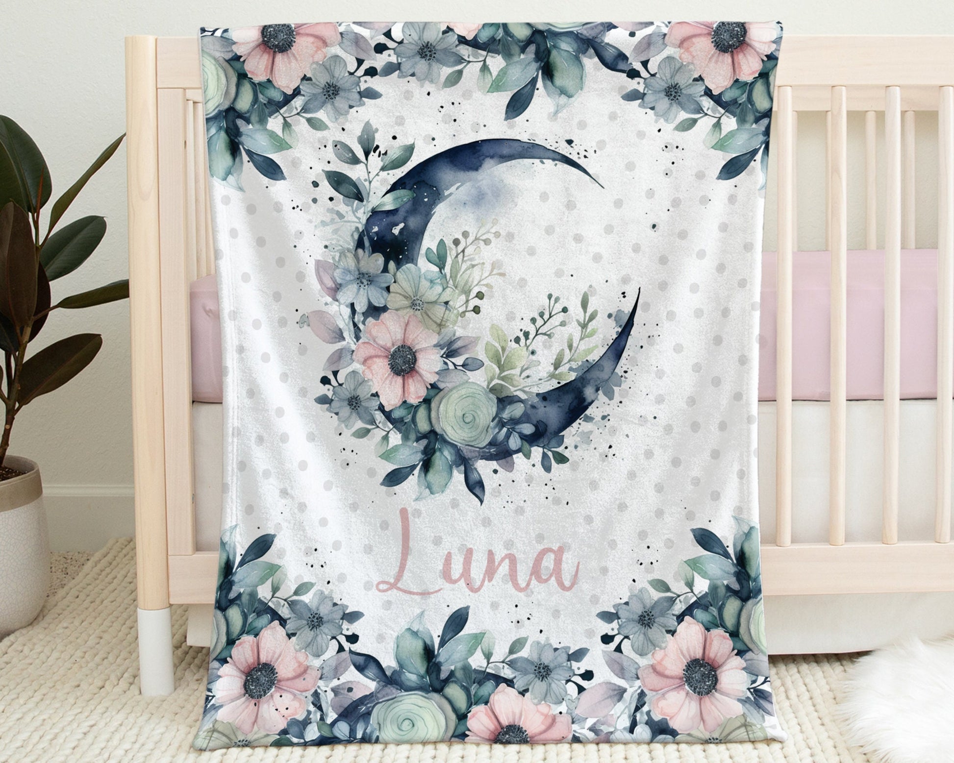 Blanket with Moon, Flowers, and Baby's Name in Pink Shown hanging on the side of a crib.
