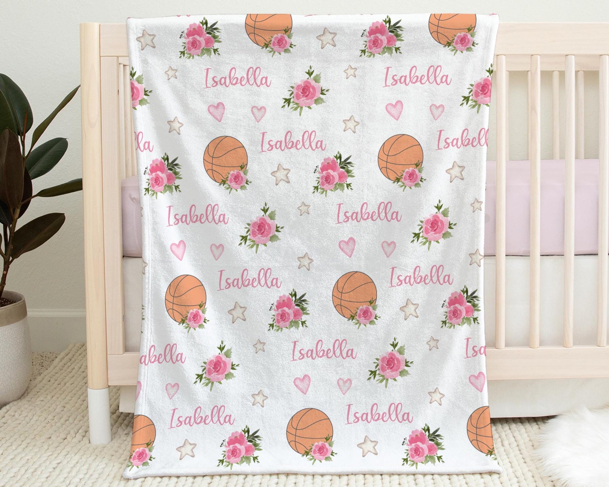 Blanket with basketballs and pink flowers that is personalized with baby's name in pink hanging on the side of a crib.