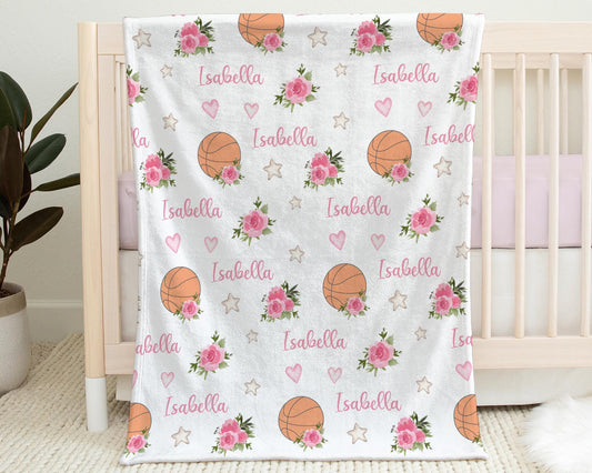 Blanket with basketballs and pink flowers that is personalized with baby's name in pink hanging on the side of a crib.