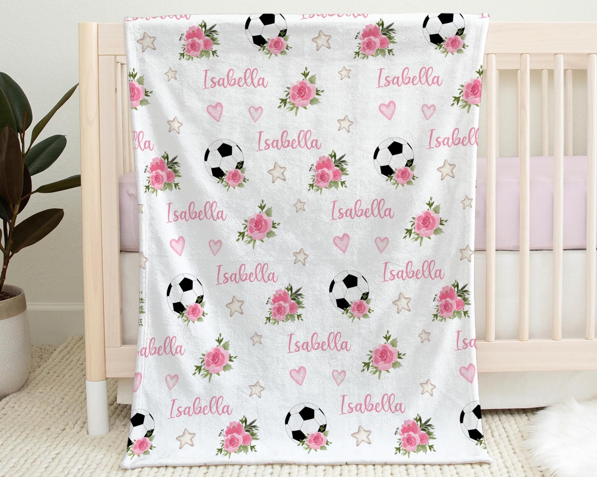 Baby Blanket with Soccer Balls, Baby's Name and Pink Flowers hanging on the side of a crib.