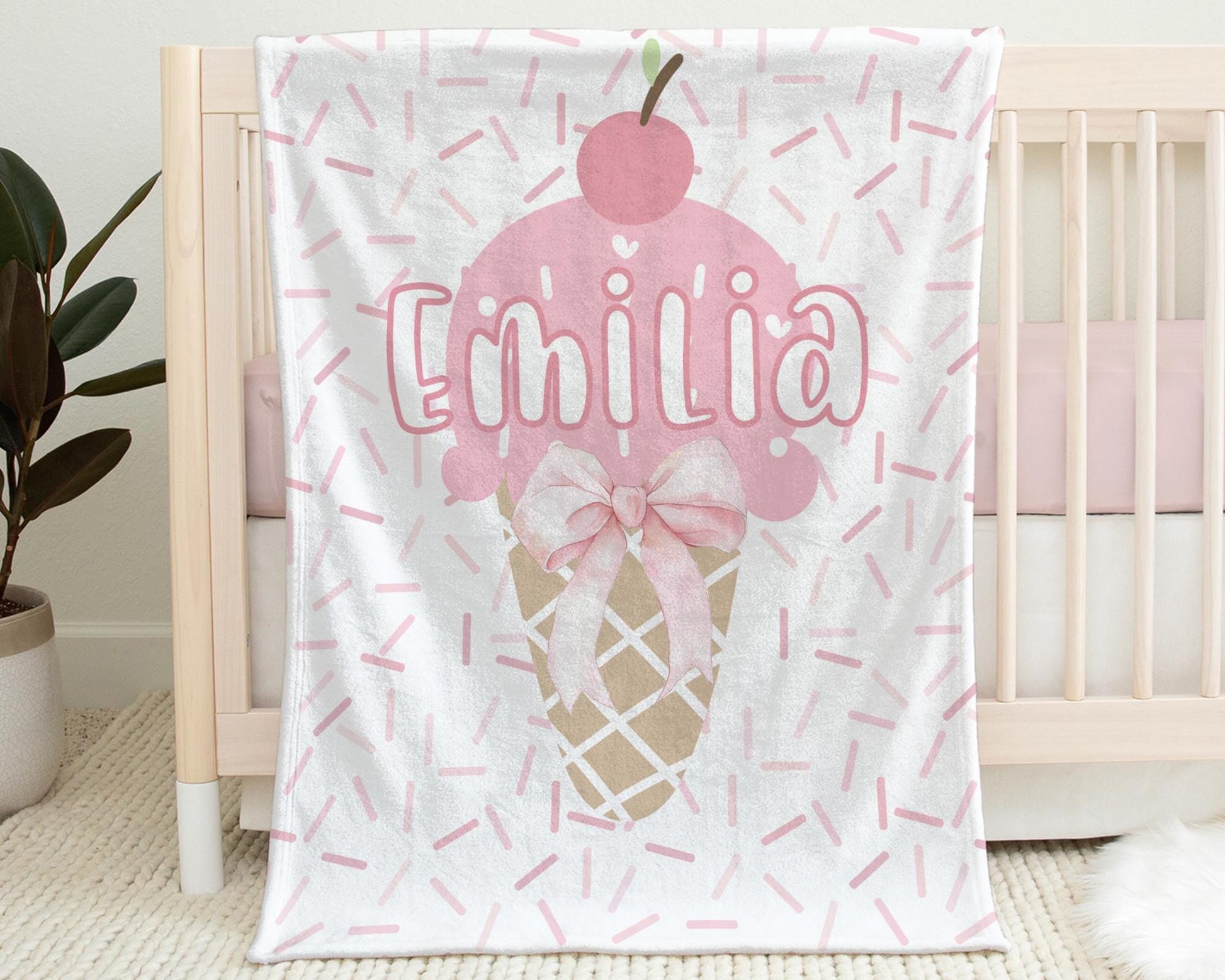Baby Blanket with Ice Cream Cone Coquette Style Bow with Sprinkles Background in Shades of Pink Pictured Hanging on the side of a crib